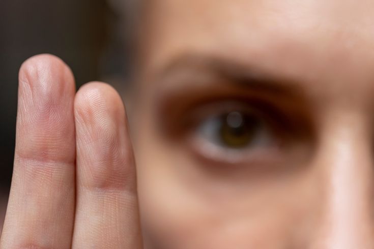 Close-up of a person's hand with fingers raised, in focus with a blurred face and eye in the background.